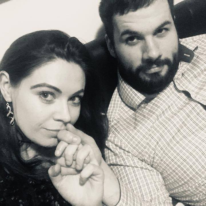 Black and white photo of a woman and man sitting closely together on a couch. The woman is holding the man's hand near her face, while the man looks directly at the camera with a calm expression.