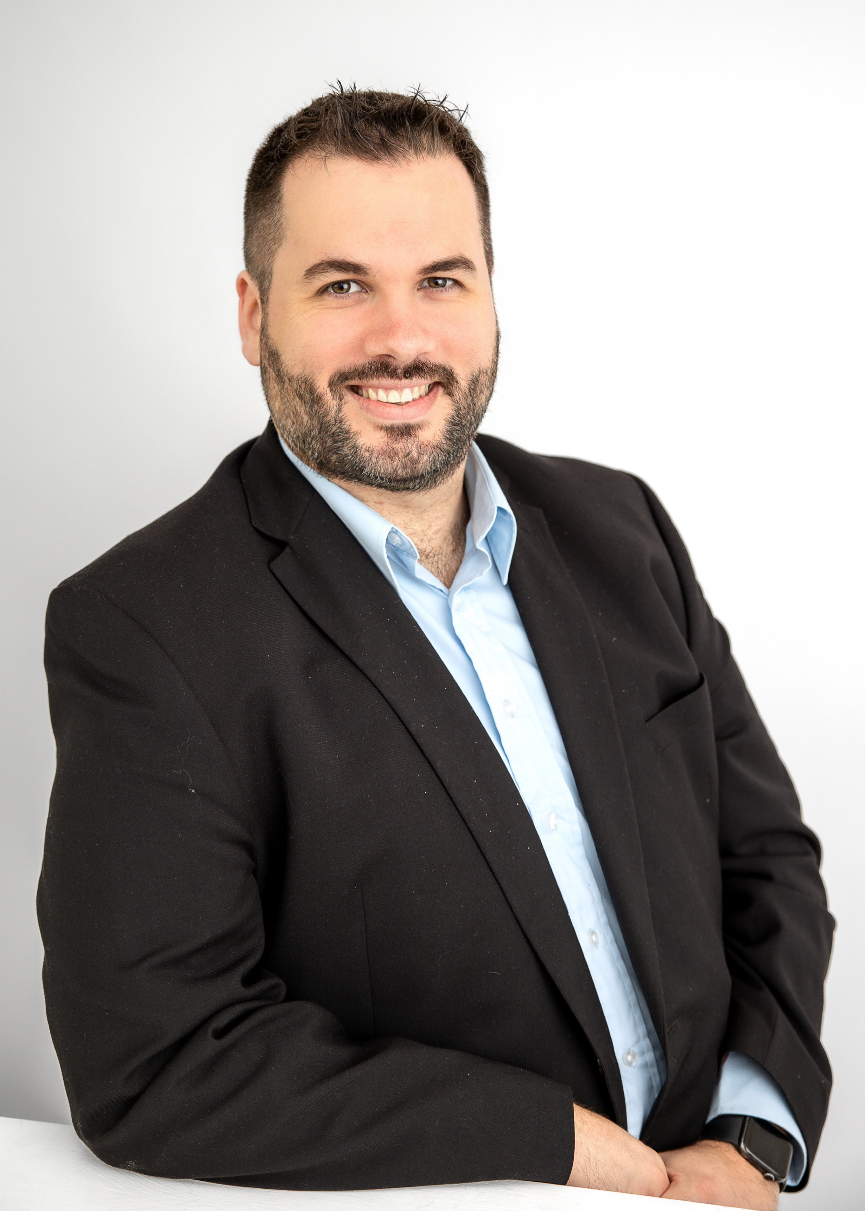 Man with short dark hair and a beard, wearing a black blazer over a light blue shirt, smiling and seated with arms crossed against a white background