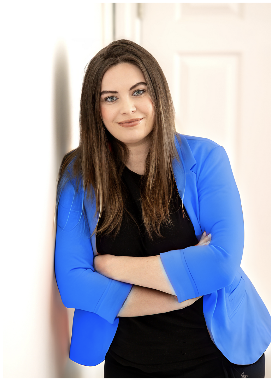 Confident woman in a blue blazer and black top, arms crossed, with a warm smile, standing indoors with a bright, blurred background