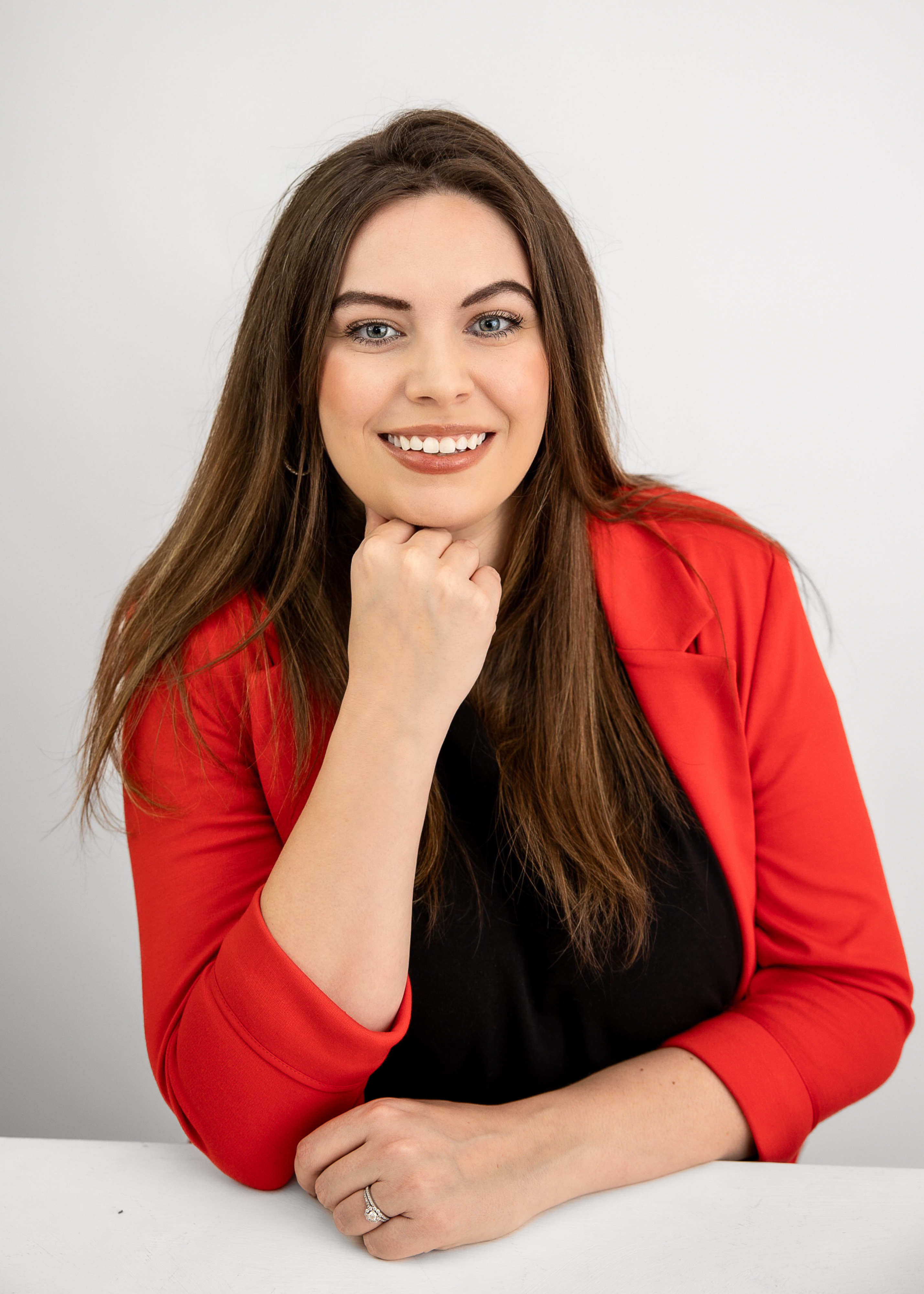 Smiling woman with long hair wearing a red blazer over a black top, resting her chin on her hand, against a grey background