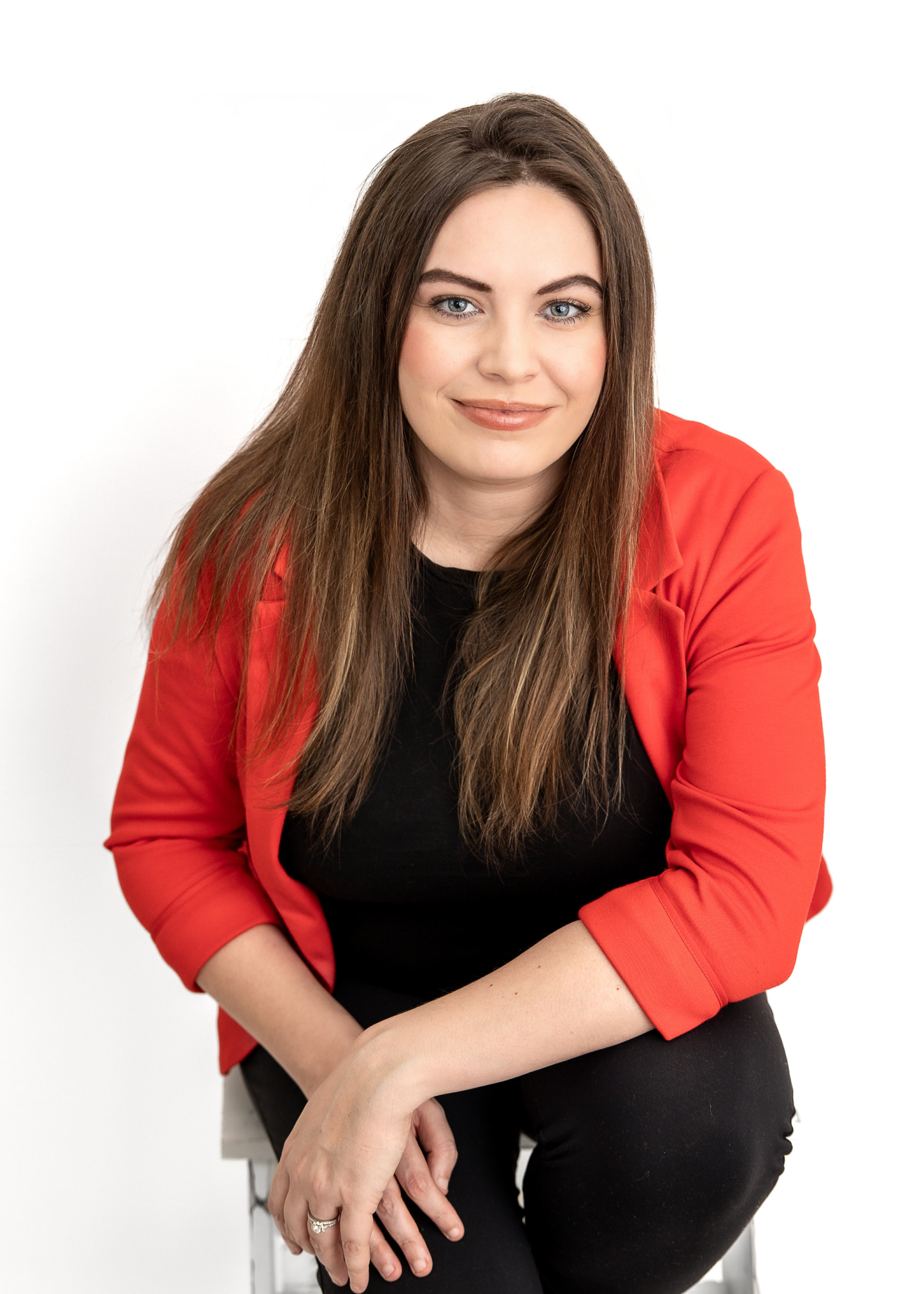 Woman with long brown hair and blue eyes, wearing a red blazer and black top, seated and smiling at the camera with a white background