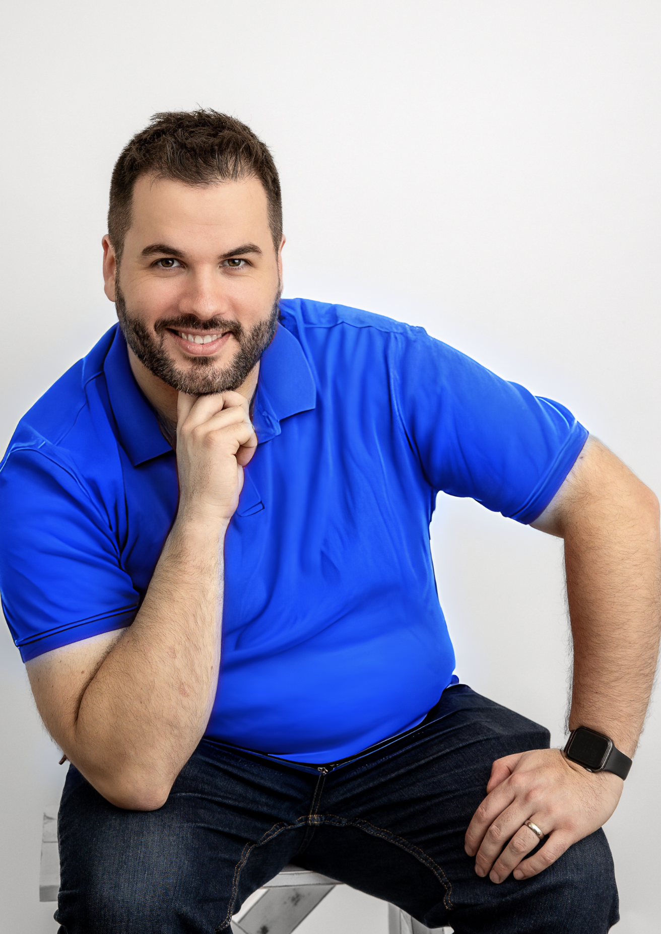 Confident man in a blue polo shirt seated with one hand under his chin, smiling at the camera, against a grey background